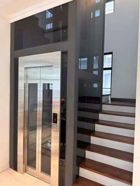 Thang máy kính khung xám lắp cạnh cầu thang gỗ biệt thự